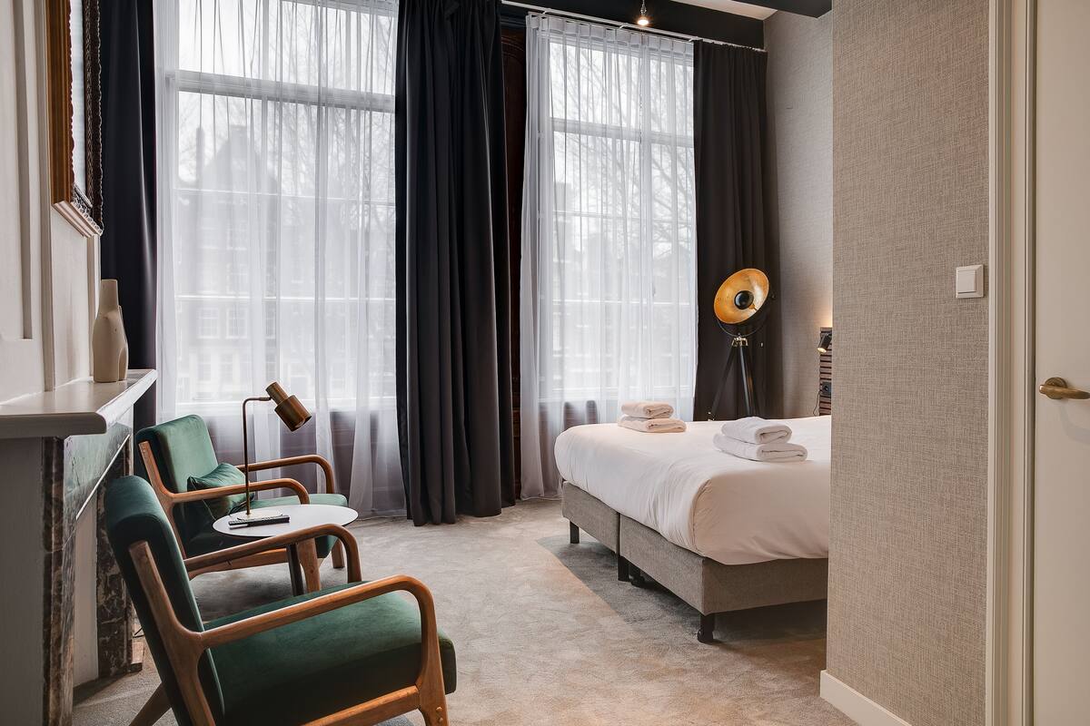 A hotel room with a bed, two green armchairs, a small table, a lamp, and a view of buildings through sheer curtains.