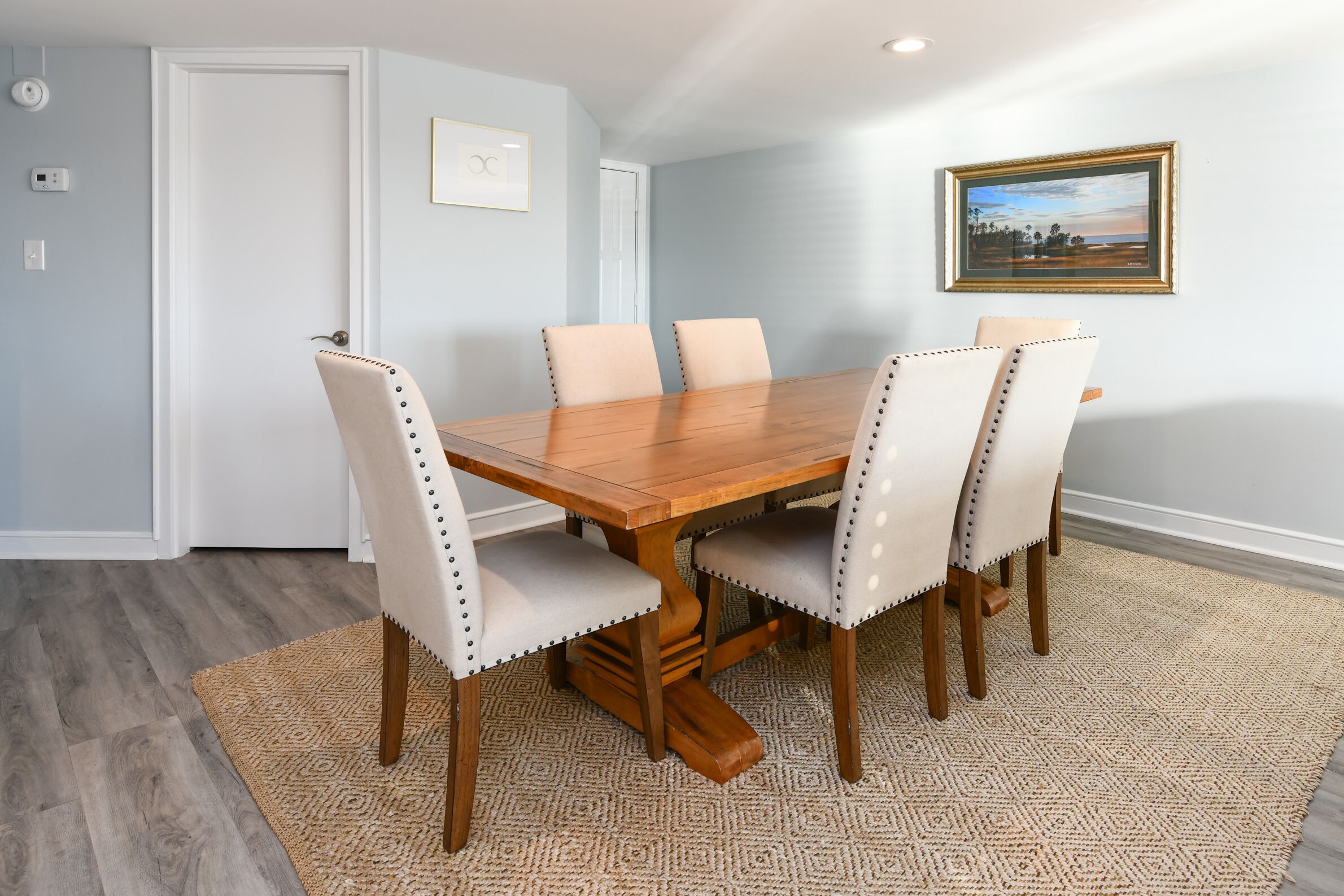 A dining area with a wooden table and six chairs, a patterned rug, and a framed picture on the wall.