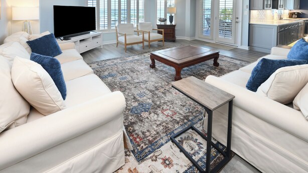 A spacious living room with a large TV, two white sofas with blue cushions, a wooden coffee table, and a patterned rug. - Inlet View 2 BR Condo! Marlin Quay 206 (Garden City)
