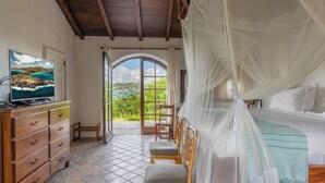 Ein Schlafzimmer mit einem Bett, einem Fernseher, einer Holzkommode, einem Sessel und Blick ins Freie.