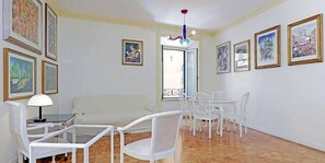 Una sala de estar con un sofá blanco, una mesa de centro de vidrio, sillas blancas y una mesa de comedor. Hay obras de arte enmarcadas en las paredes y un balcón con barandilla.