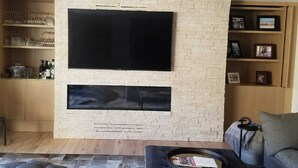 A modern living room with a large flat-screen TV mounted on a brick wall, a fireplace below, and a cozy seating area with a sofa and armchairs.
