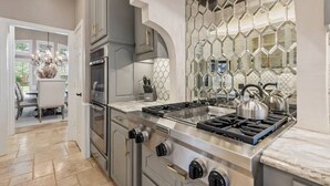 A modern kitchen with a stainless steel stove, a microwave, and a built-in oven. There is a hexagonal tile backsplash and a dining area visible through an open door. - Luxurious Home-Resort Style Pool & Game Room (Plano)
