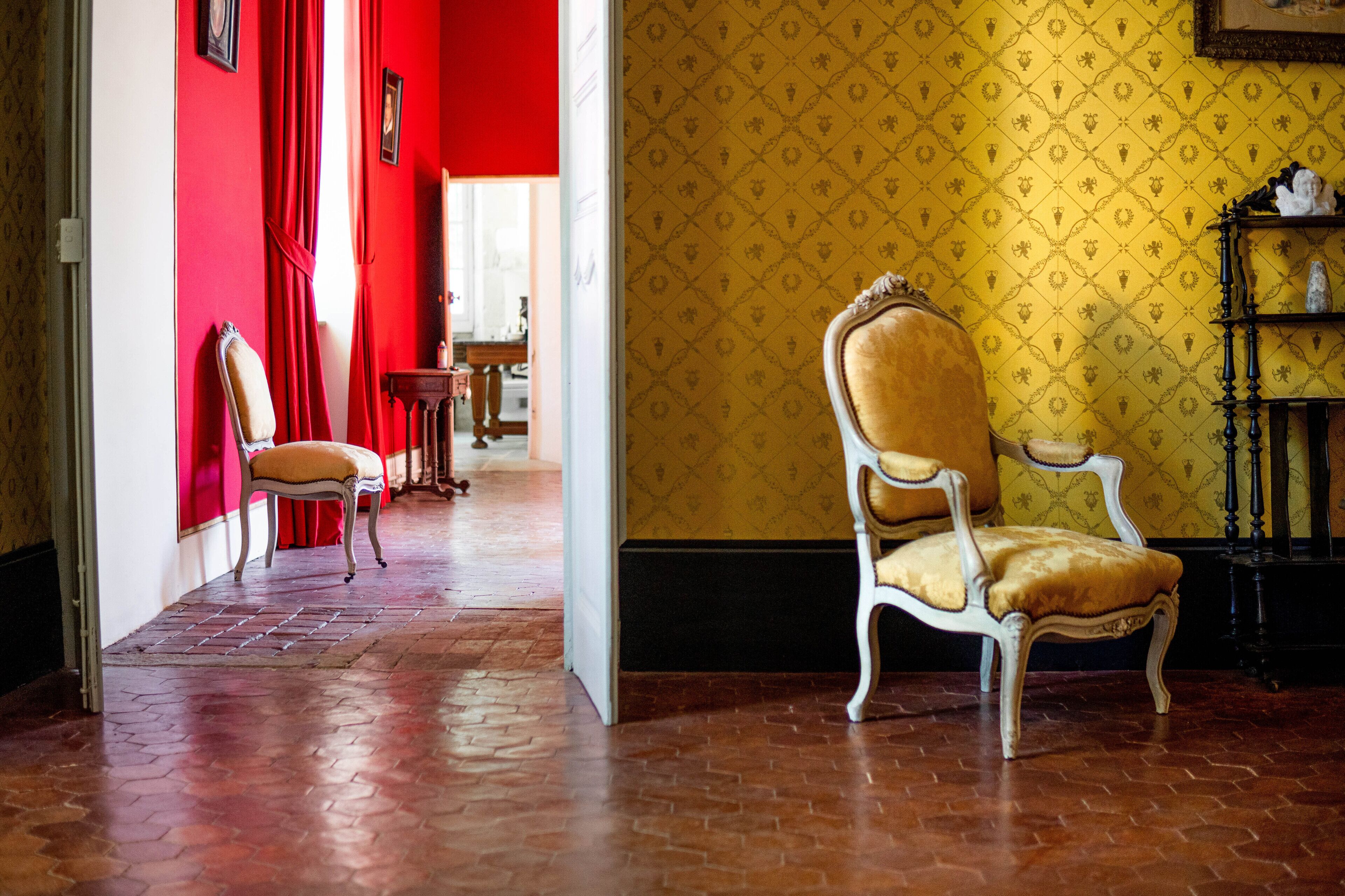 A hallway with a red wall, a yellow patterned wall, and a brick floor. There are two chairs and a table in the hallway.