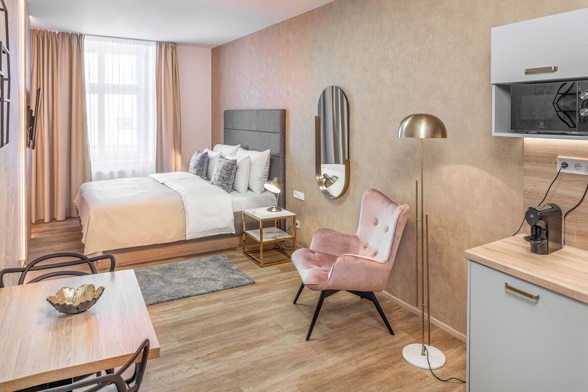 A modern hotel room with a bed, bedside table, chair, desk, and a small kitchenette.