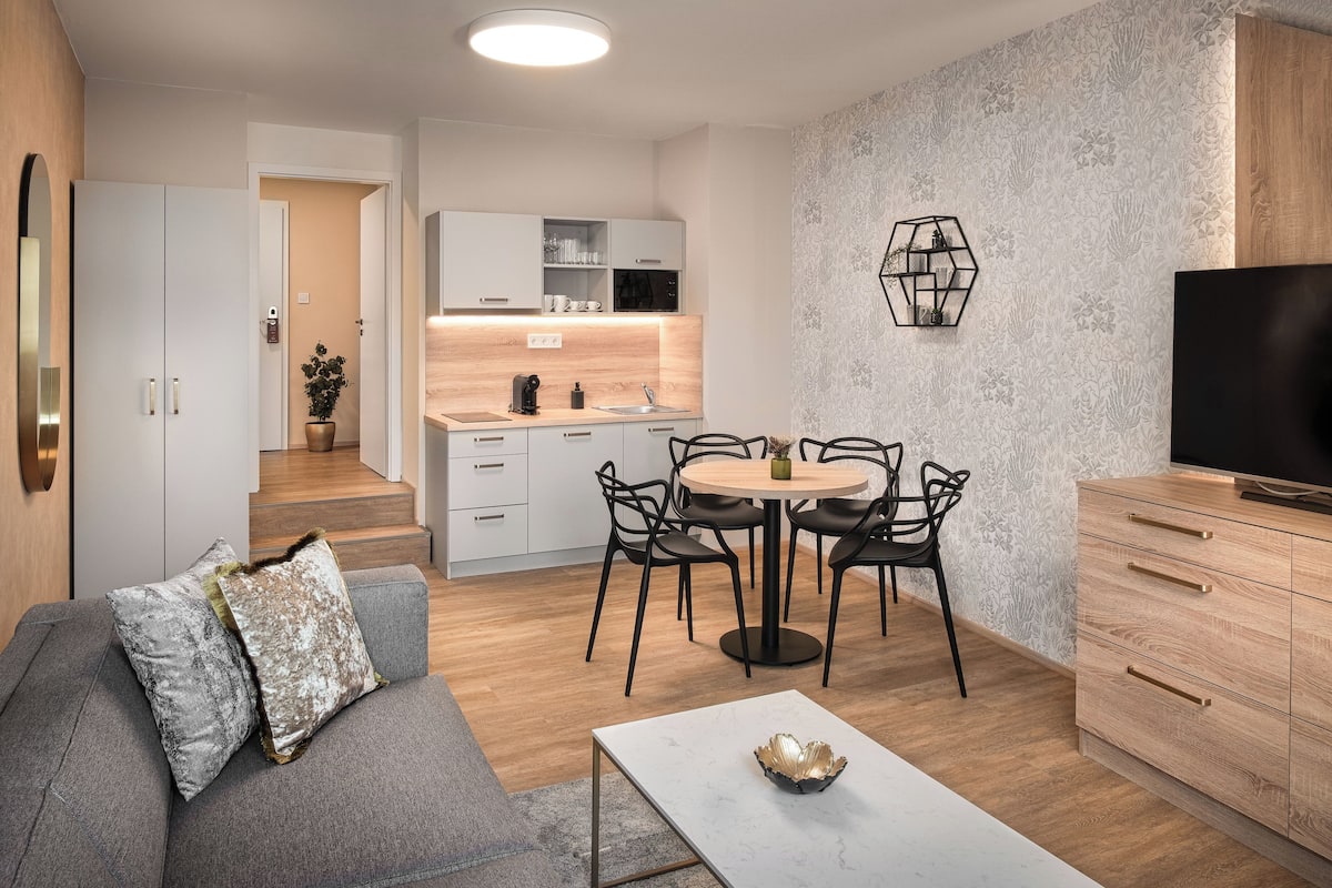 A modern living room with a sofa, coffee table, and a dining area with chairs and a table. There is a kitchen with cabinets and a countertop. A TV is mounted on the wall, and there is a staircase leading to another level.