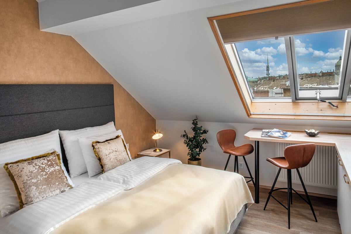 A modern bedroom with a sloped ceiling, a bed with white linens, a desk with two chairs, and a view of a cityscape through large windows.