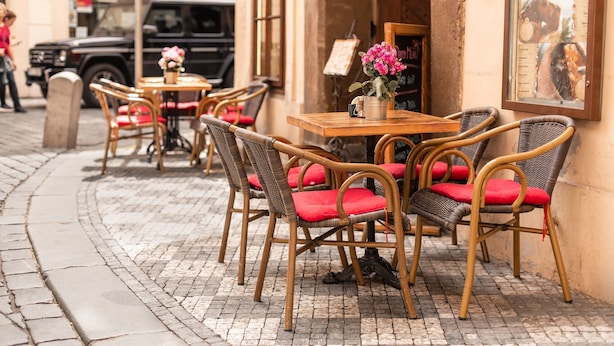 Espace extérieur aménagé avec des chaises en osier et des coussins rouges, une table en bois et un panneau affichant le menu, le tout installé sur le mur extérieur d’un bâtiment.