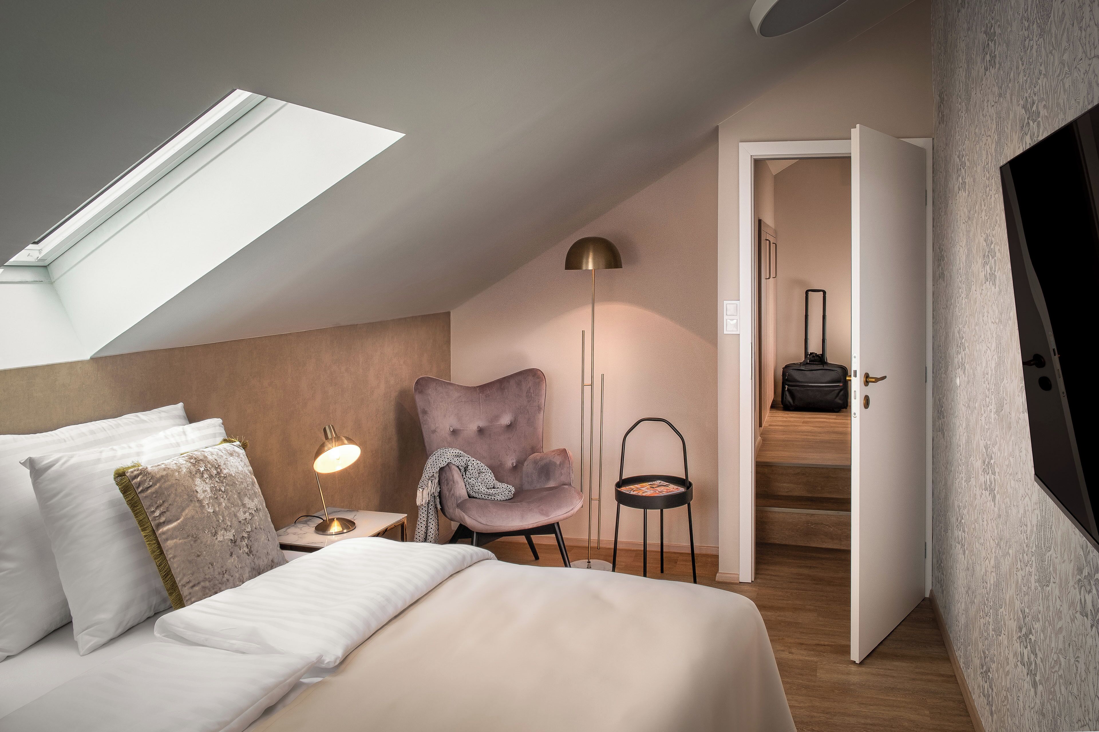 Een slaapkamer met dakraam, een bed met kussens, een stoel, een vloerlamp en een koffer op de vloer.