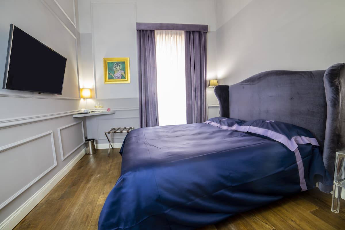 Un dormitorio con una cama grande, un televisor de pantalla plana montado en la pared, una mesita de noche con una lámpara y una ventana con cortinas.
