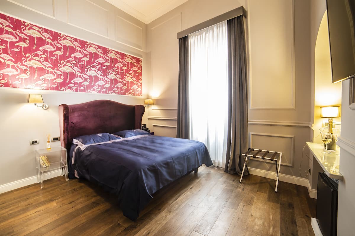 Habitación de hotel con una cama grande, pared floral roja, suelo de madera y televisor a la derecha.