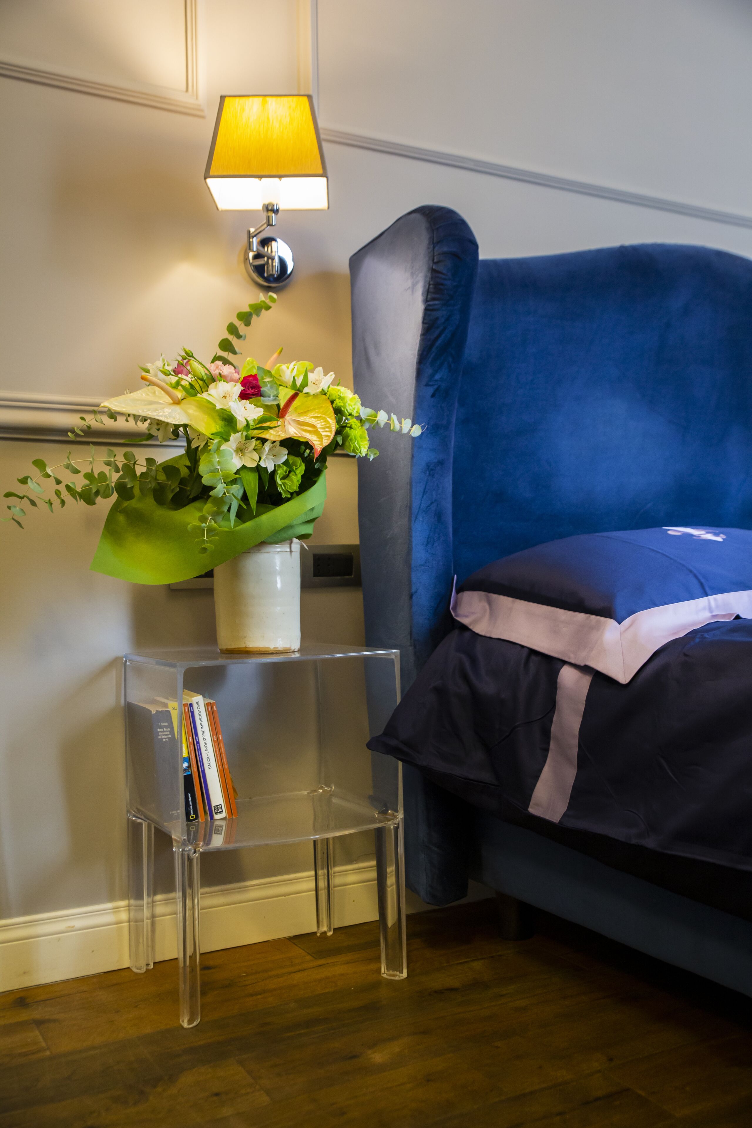Quarto com sofá azul, mesinha lateral transparente com livros, um vaso com flores e luminária na parede.