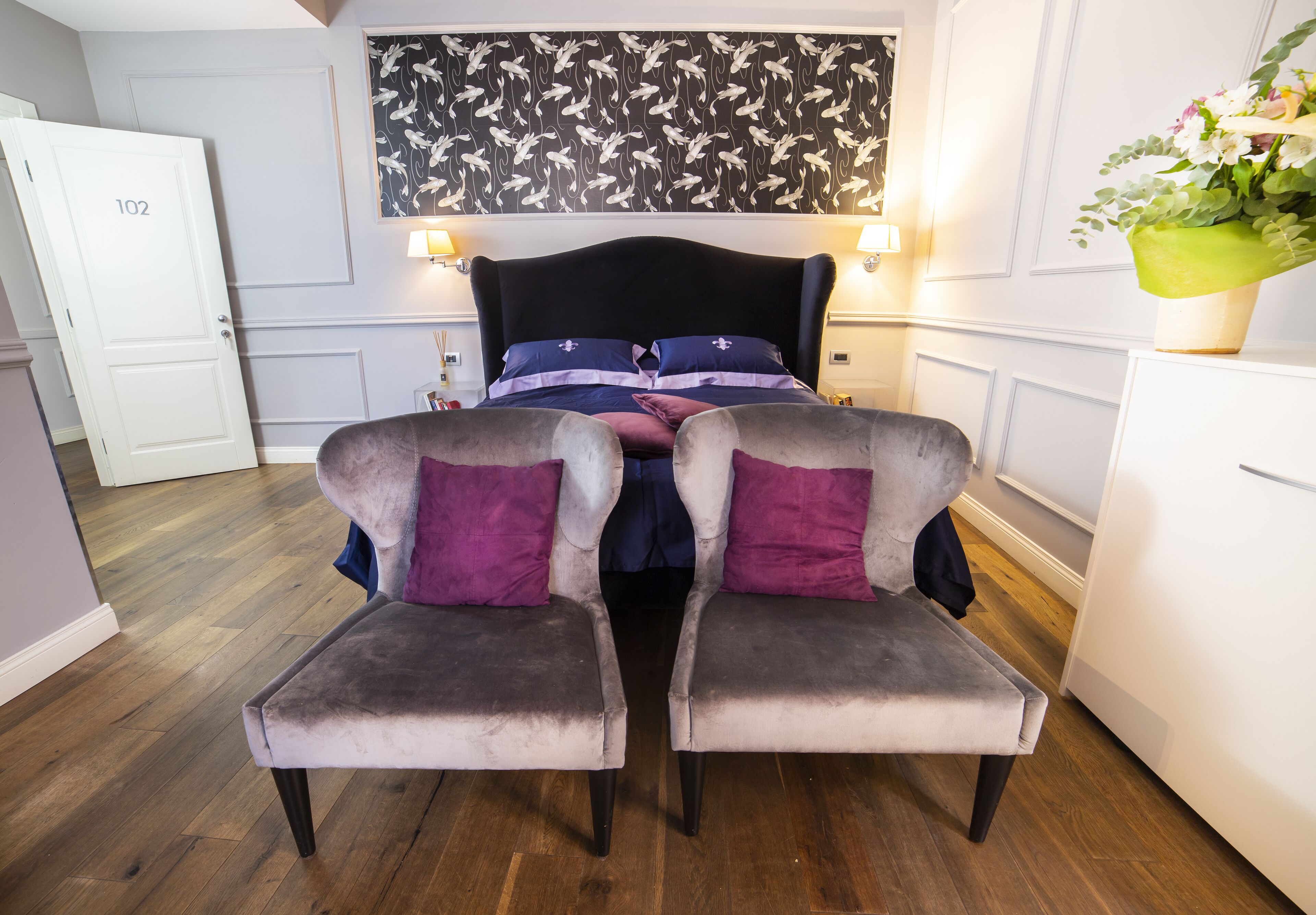 Un dormitorio con una cama, dos sillones, un aparador y una puerta con la leyenda '102'.
