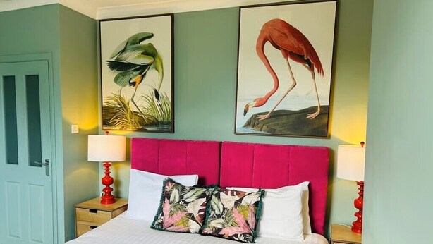 Una habitación de hotel con una cama grande, dos cuadros de aves colgados en la pared, un cabecero rojo y una puerta verde azulada.