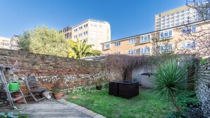 Un patio trasero con terraza de madera, pared de ladrillo y un pequeño jardín con plantas en macetas y una palmera.