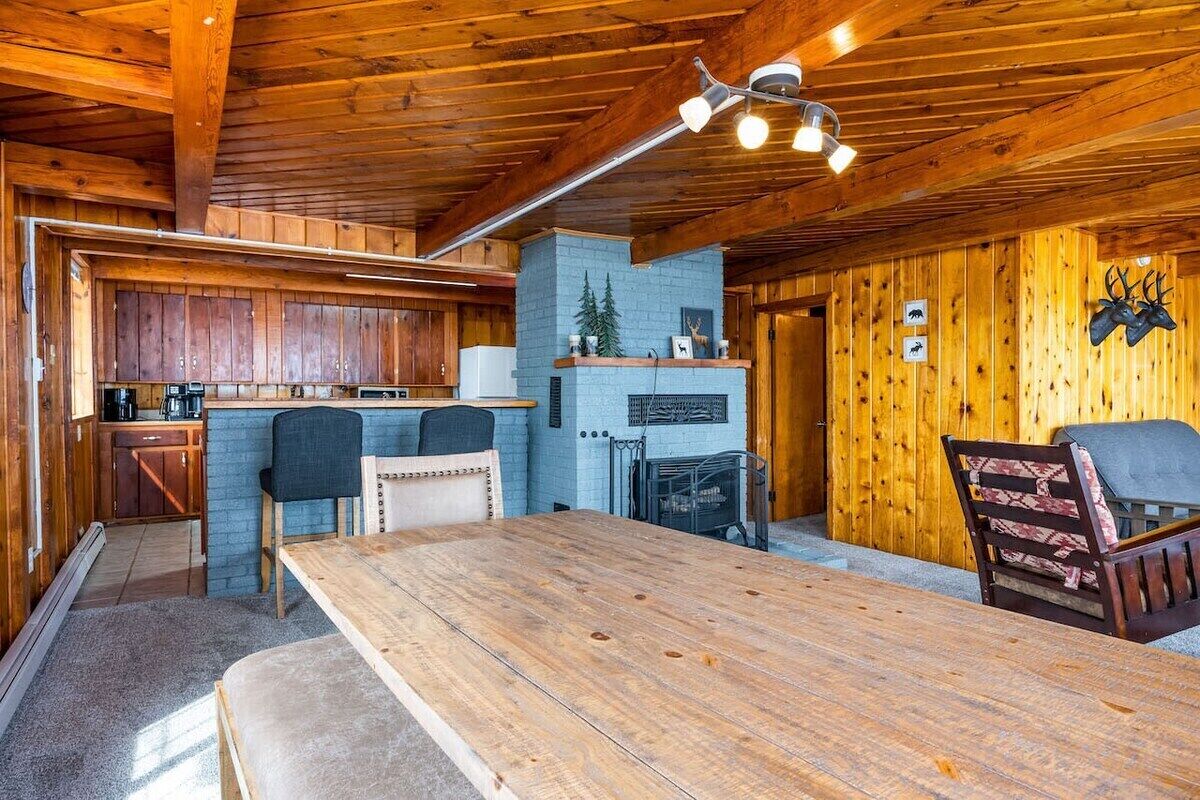 A rustic cabin interior with a wooden dining table, two chairs, a fireplace, and a kitchen area.
