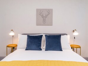 Un lit bien fait, recouvert d’une couette blanche et bleue, avec deux oreillers bleus et un plaid jaune. Deux tables de chevet, chacune surmontée d’une lampe, se trouvent de part et d’autre du lit.