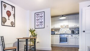 Une cuisine moderne avec des armoires blanches, un dosseret carrelé et une cuisinière. On y trouve un coin repas avec une table et des chaises en bois, ainsi qu’une plante posée sur la table.