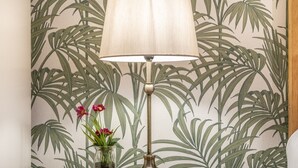 Une lampe avec un abat-jour beige, posée sur une table en bois, devant un mur orné d’un motif de feuilles tropicales.