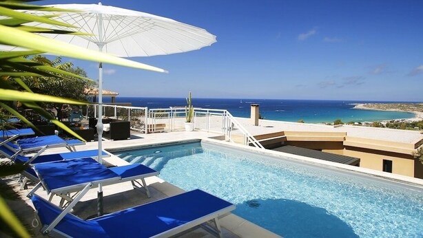 Un espace piscine avec des chaises longues bleues et un parasol blanc, offrant une vue sur la côte.