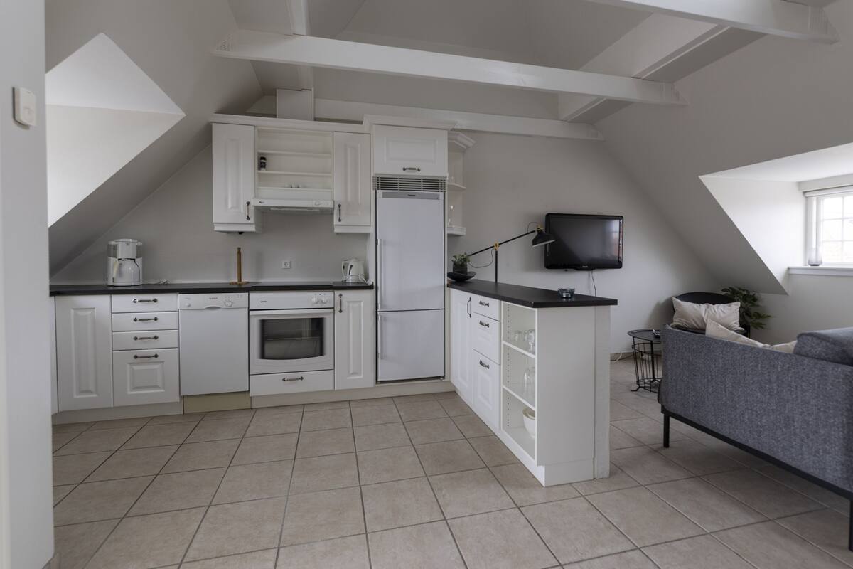 A modern kitchen with white cabinets, a built-in oven, and a refrigerator. There is a gray sofa and a TV mounted on the wall.