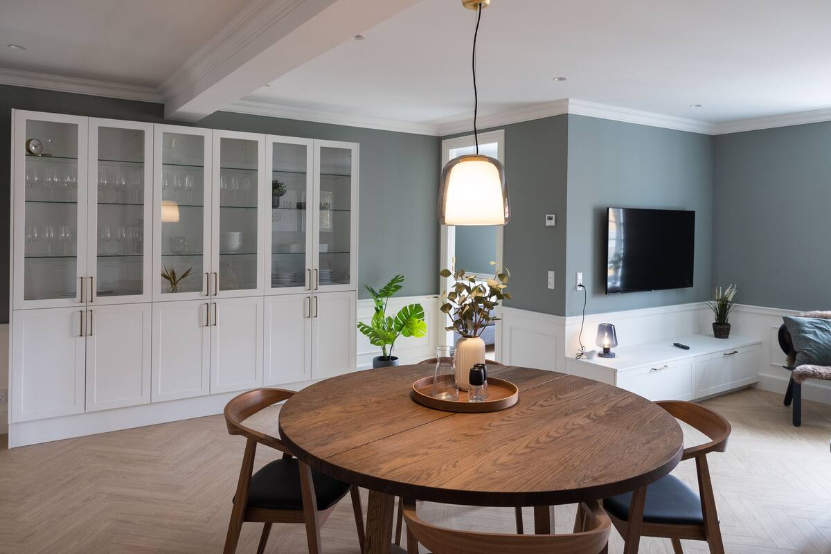 A modern kitchen with white cabinets, a round wooden dining table, and a built-in TV.