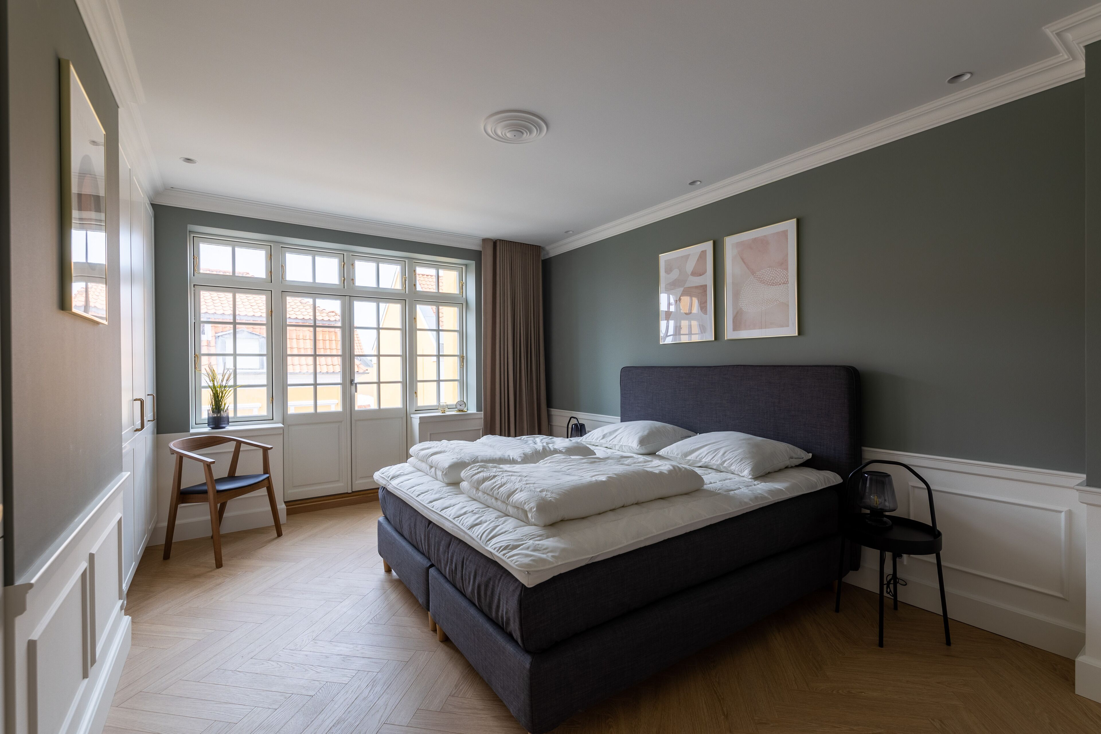 Un dormitorio con una cama grande, una ventana con cortinas, una silla y obras de arte en la pared.