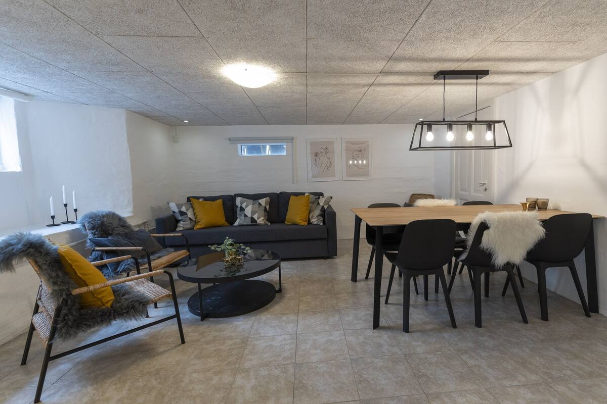 A modern living room with a grey sofa, black and yellow chairs, a wooden dining table, and a hanging light fixture.
