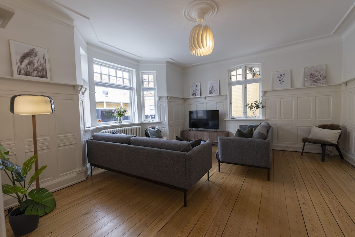 A spacious living room with a large sofa, a smaller armchair, a TV, and a potted plant.