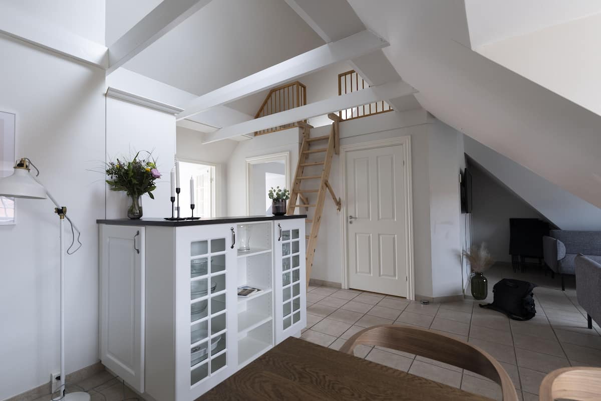 A spacious loft with a high ceiling, a wooden staircase, and a kitchen area with white cabinetry and a dining table.