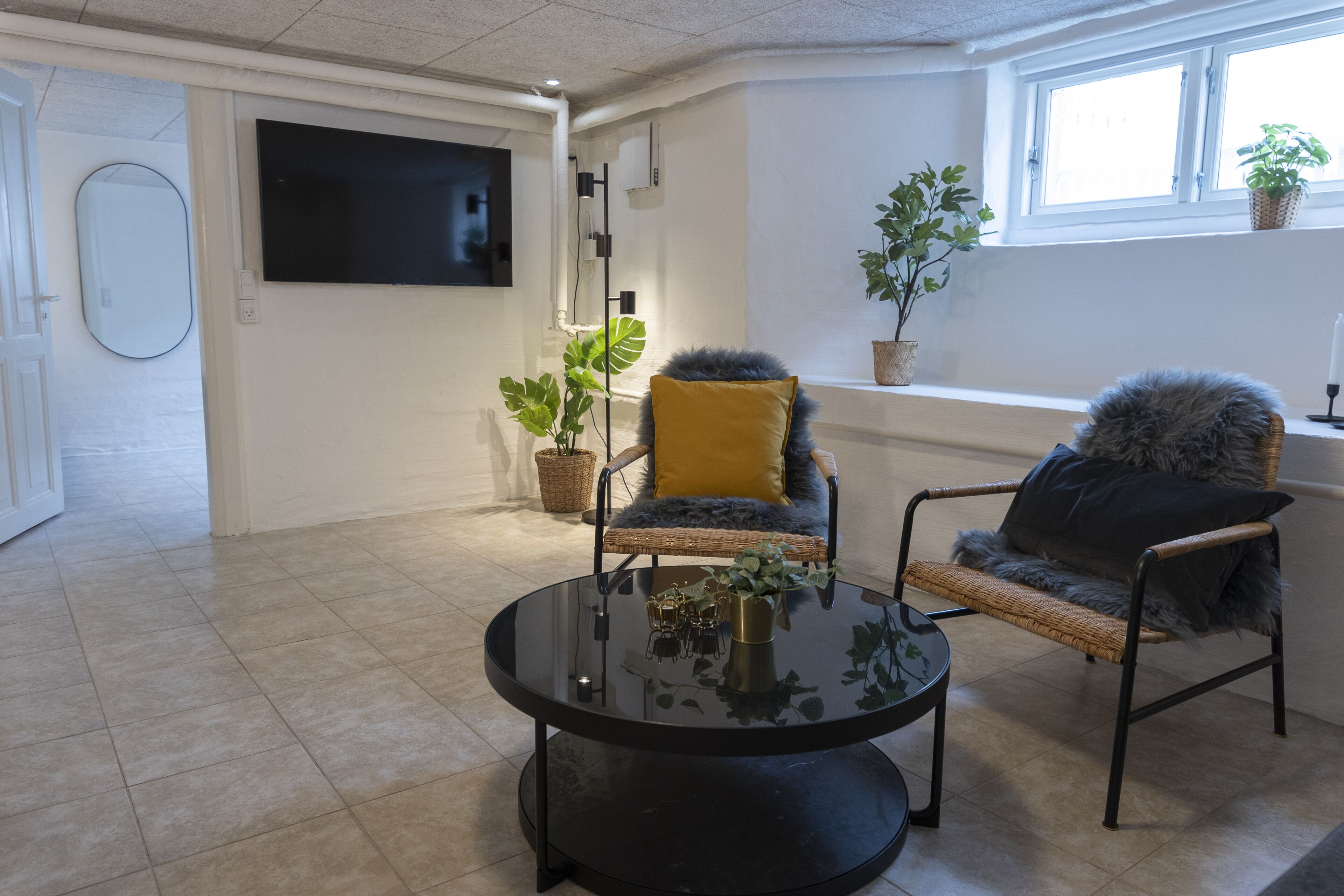 Un salon moderne avec une table ronde noire, deux fauteuils en osier avec des coussins, un grand téléviseur fixé au mur et des plantes en pot près de la fenêtre.