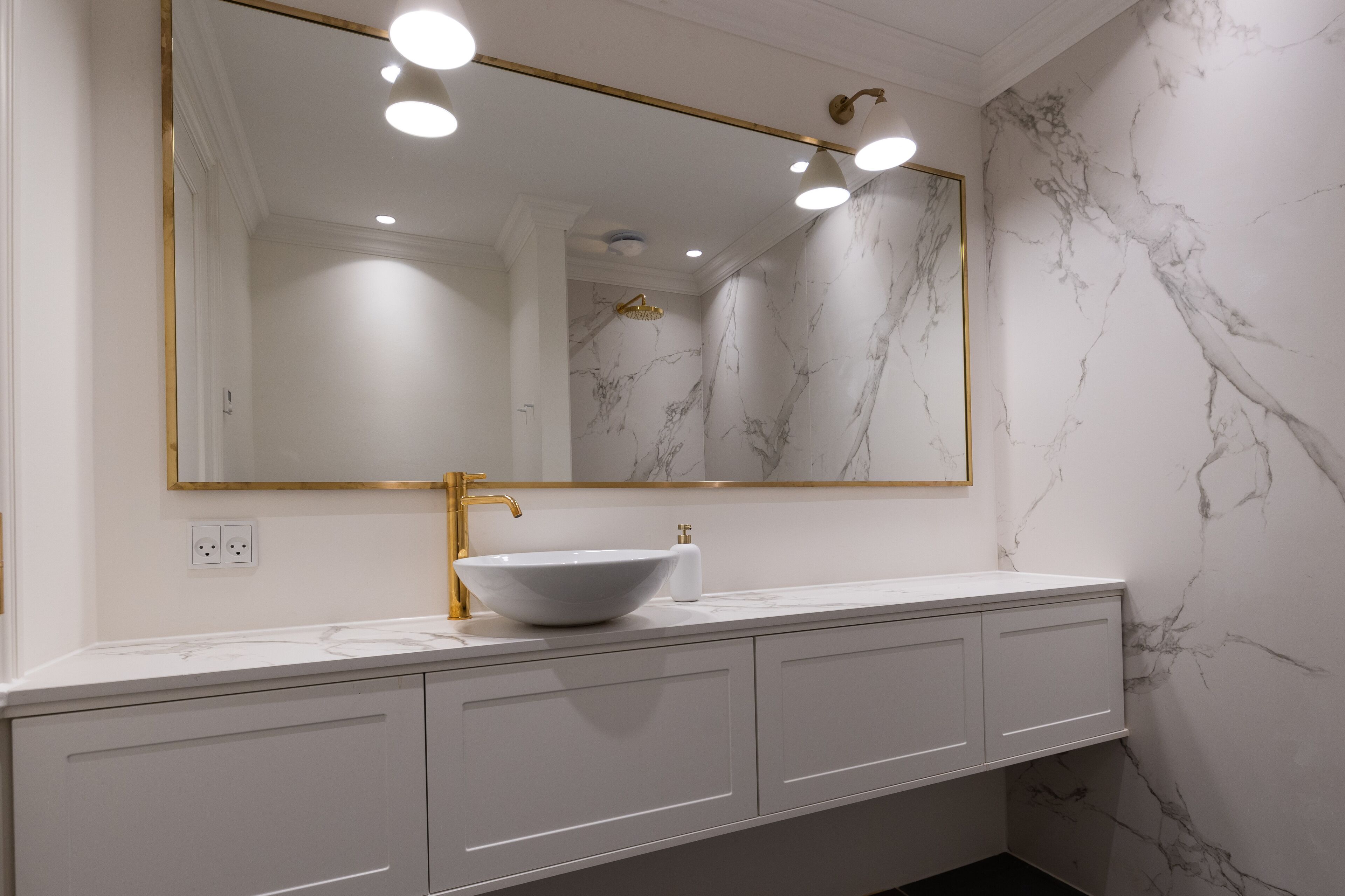 A bathroom with a white marble wall, a gold-framed mirror, a white sink, and a gold faucet.
