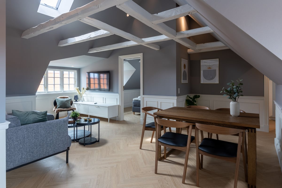 A modern loft apartment with exposed wooden beams, a dining area with wooden chairs, a grey sofa, and a kitchen with white cabinets.