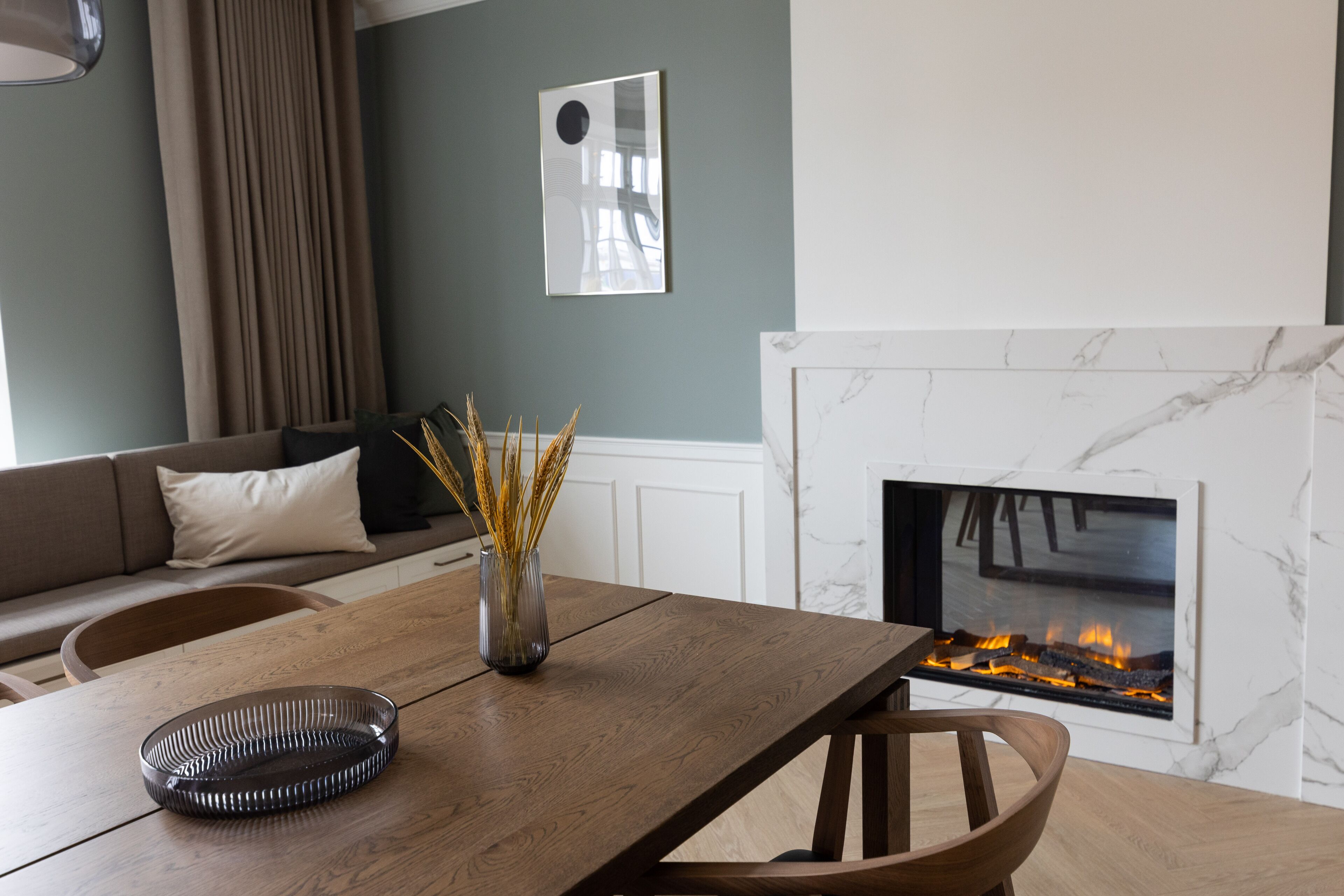 Un comedor con una mesa de madera, sillas, un florero con plantas secas y una chimenea encendida.