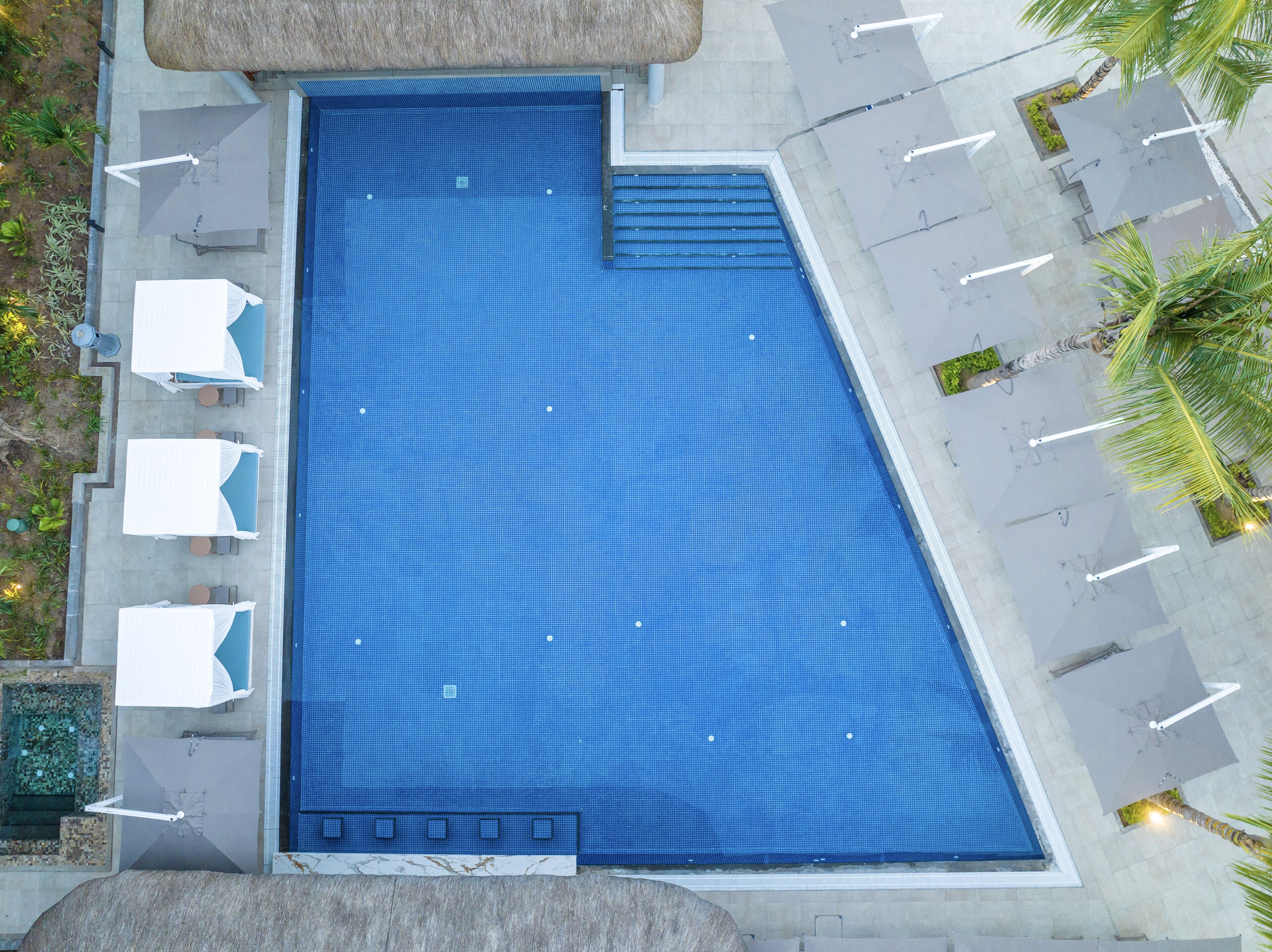 Una amplia piscina rectangular con superficie de azulejos azules, rodeada de sillas de descanso y sombrillas.