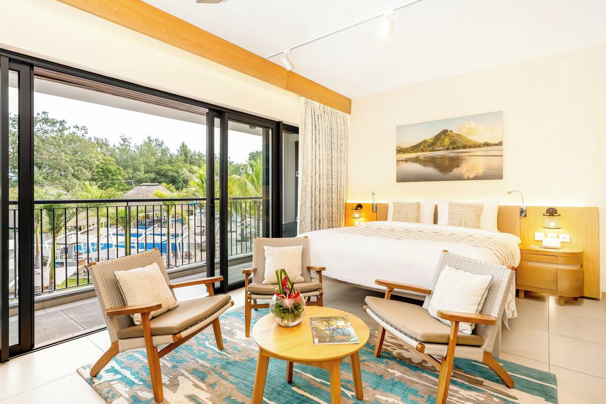 Une chambre d’hôtel avec un grand lit, deux fauteuils, une petite table avec un vase, et une vue sur une piscine et un espace verdoyant.