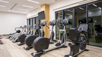 A row of stationary bikes and elliptical machines in a gym with large windows and a wooden floor.
