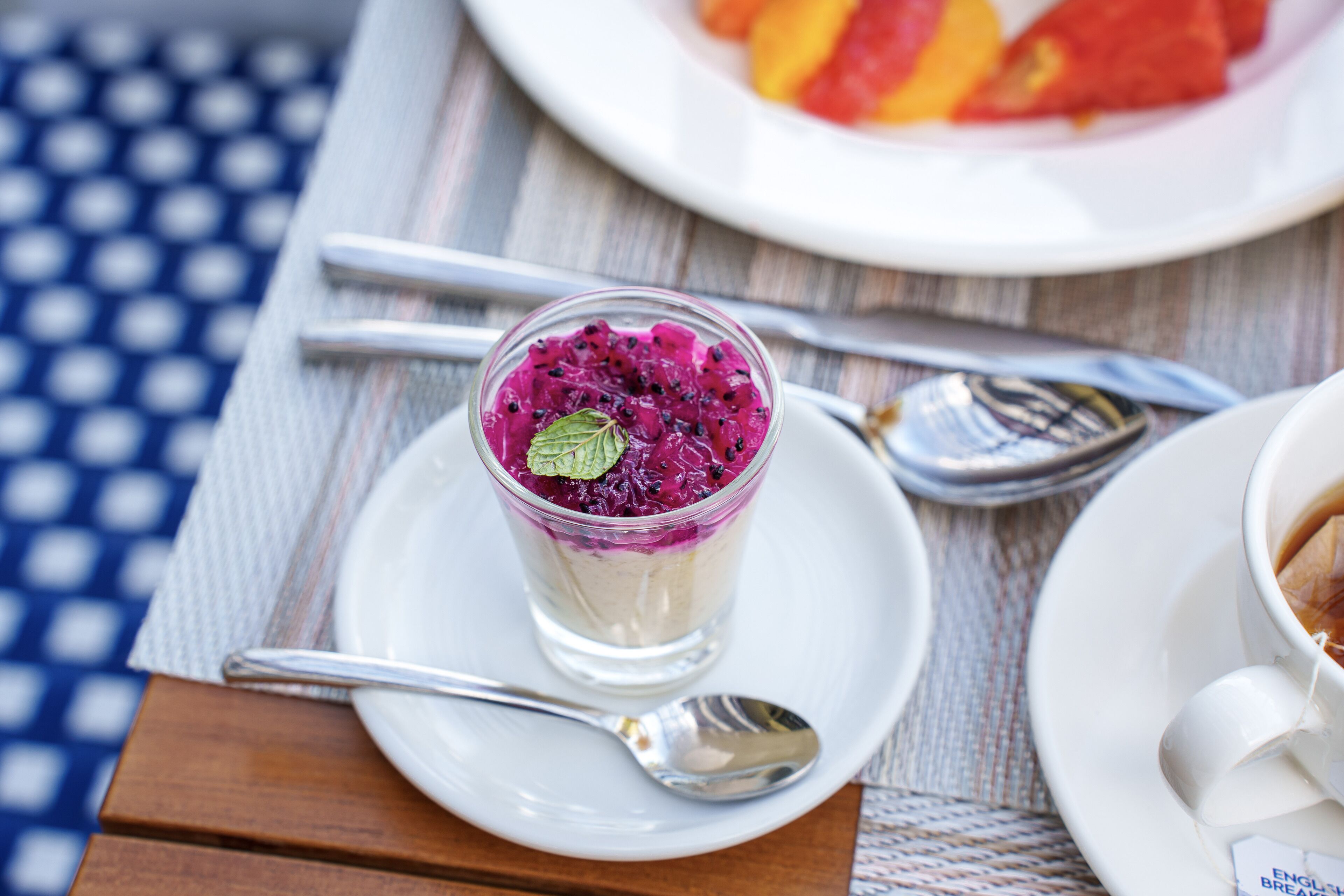 Un postre en vaso sobre un plato blanco con una cuchara al lado, una taza de café en su platito y un plato de fruta al fondo.