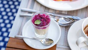 Un postre en vaso sobre un plato blanco con una cuchara al lado, una taza de café en su platito y un plato de fruta al fondo.