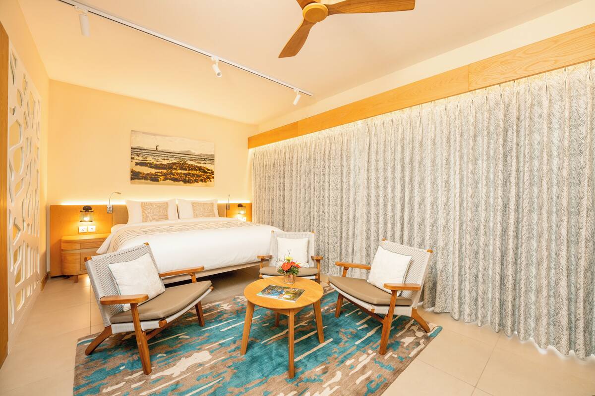 Une chambre d’hôtel avec un lit, deux fauteuils, une petite table sur laquelle est posé un vase de fleurs et un ventilateur de plafond.