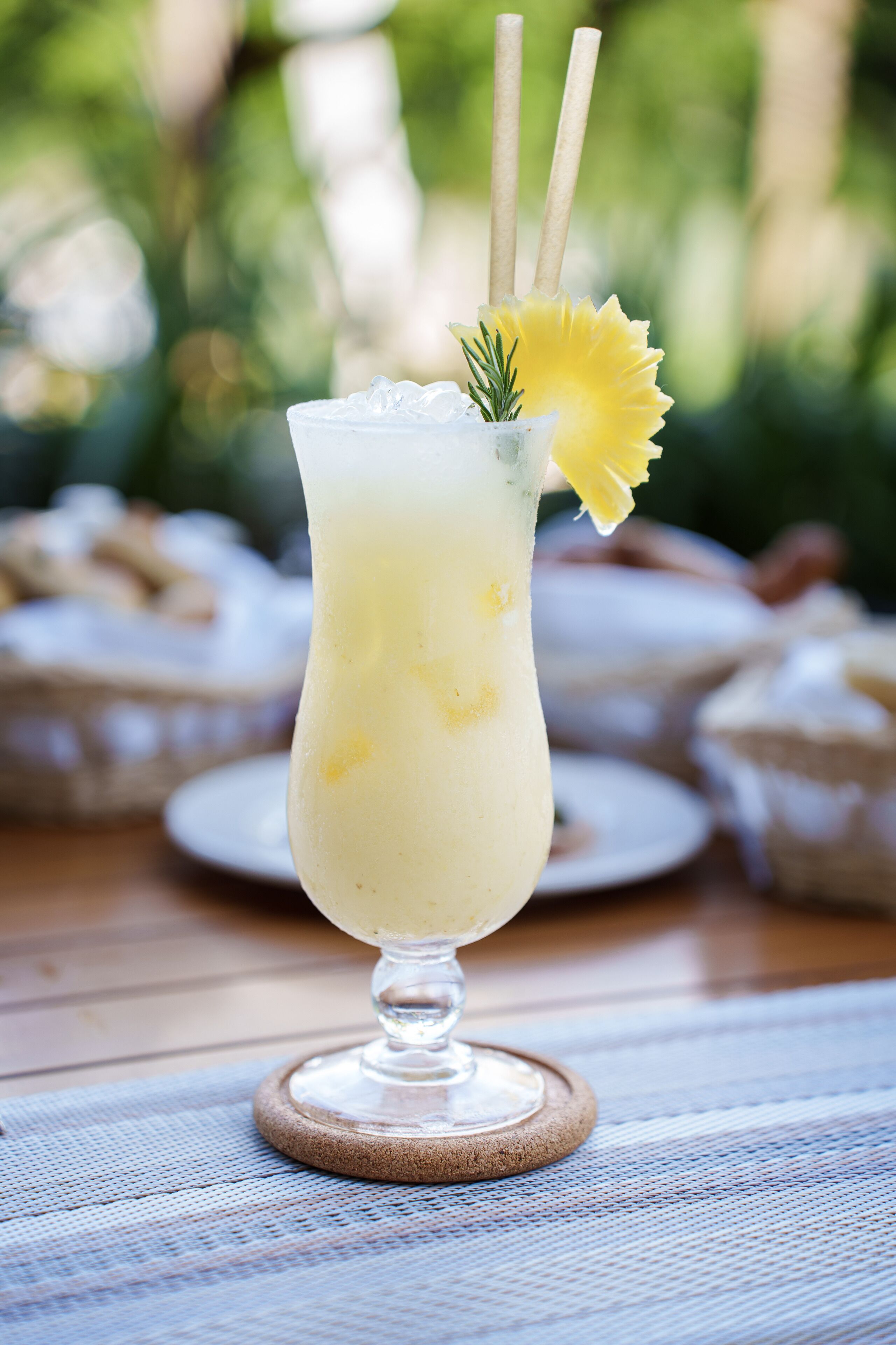 A tall glass with a yellow drink, garnished with a flower and a sprig, on a wooden coaster.