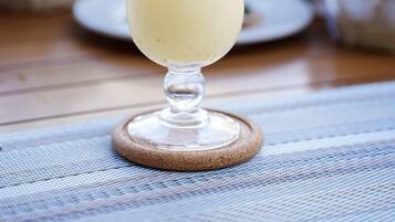 A tall glass with a yellow drink, garnished with a flower and a sprig, on a wooden coaster.