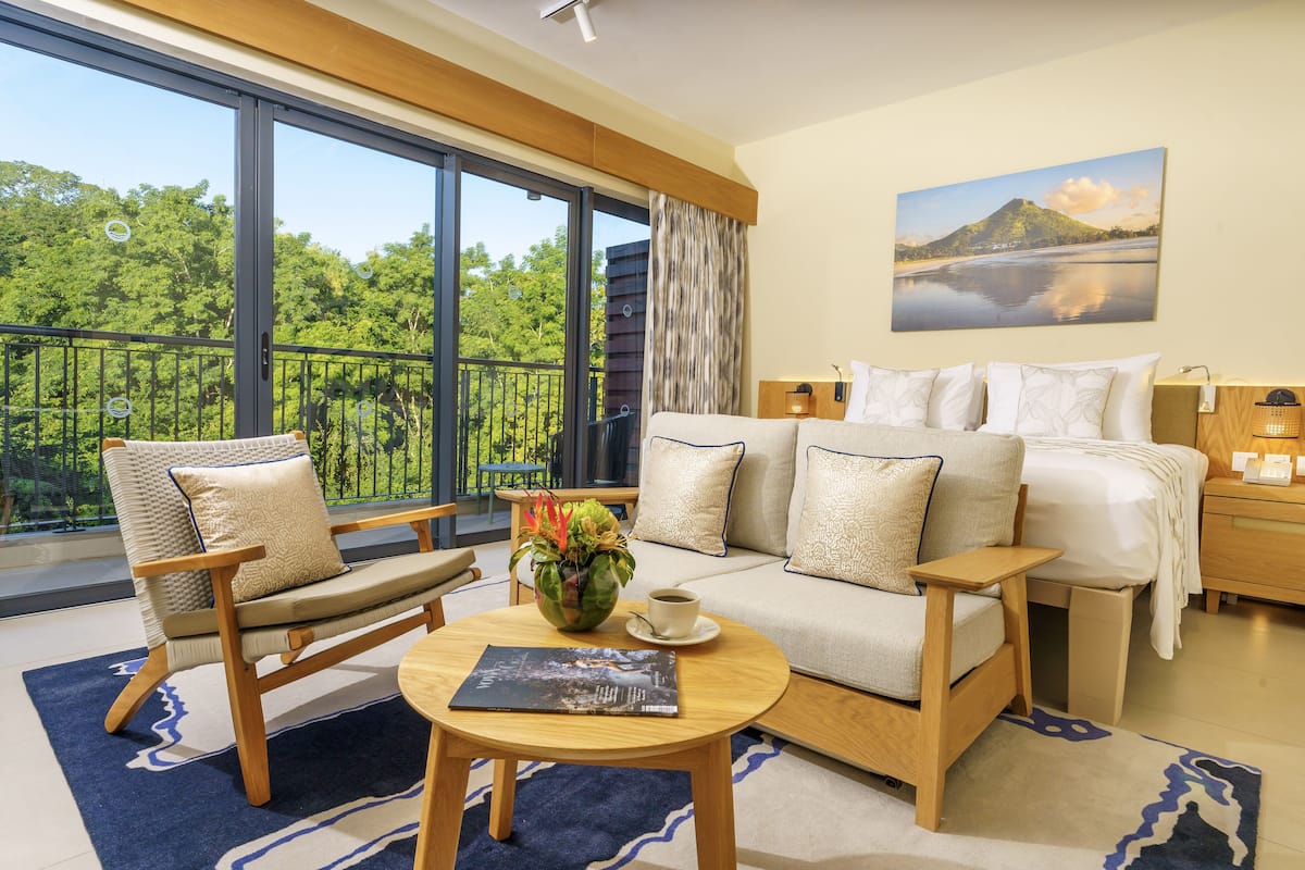 Une chambre d’hôtel moderne dotée d’un grand balcon, d’un canapé, d’un fauteuil, d’une table de chevet et d’un tableau représentant un paysage.