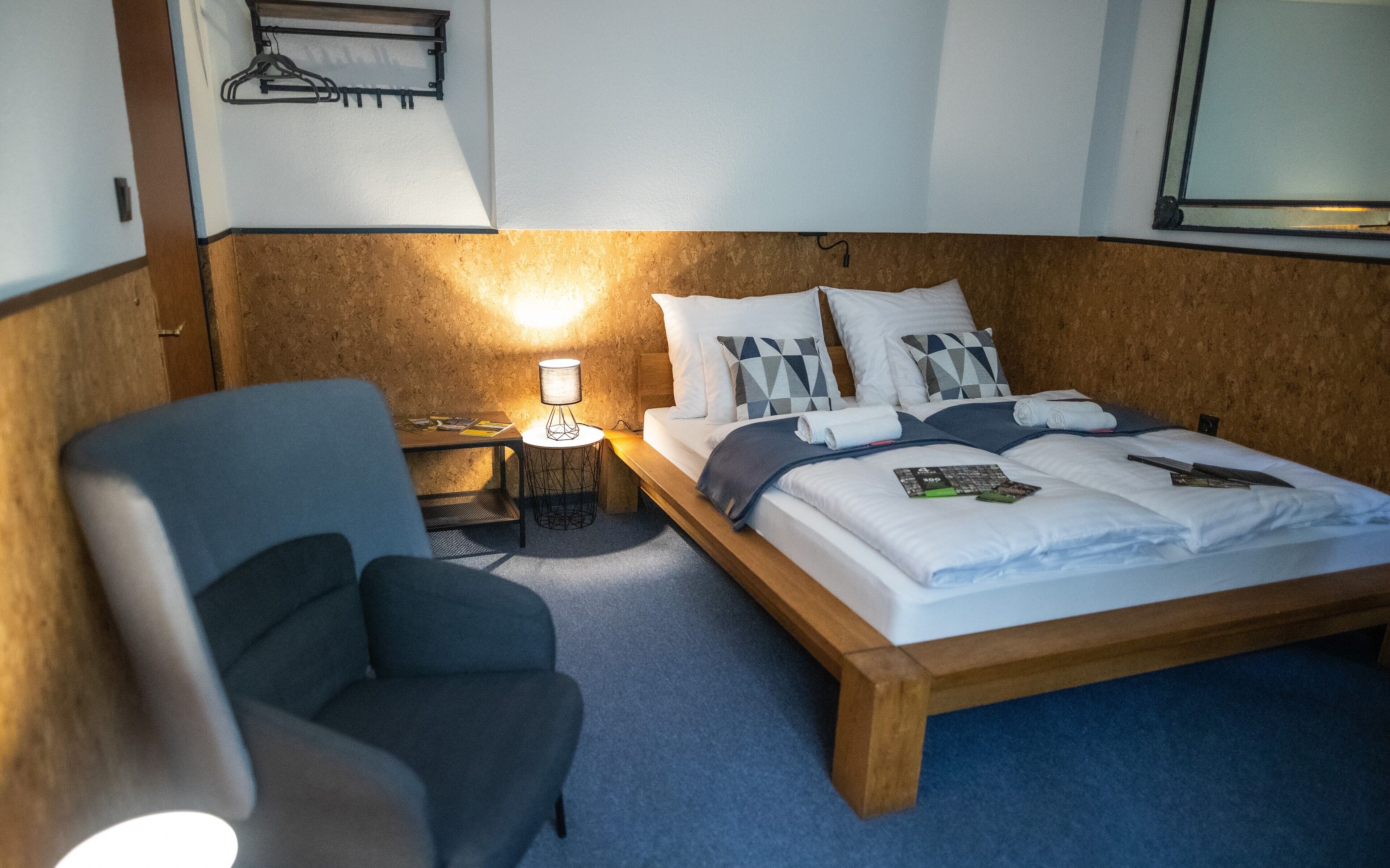 Ein Hotelzimmer mit einem hölzernen Bett, einem Sessel, einem kleinen Tisch mit Lampe und einem Wandhaken zur Kleideraufbewahrung.