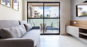 A modern living room with a grey sofa, a TV mounted on the wall, and a balcony with a chair. - Apart térreo no Porto Blue por Carpediem (Ipojuca)