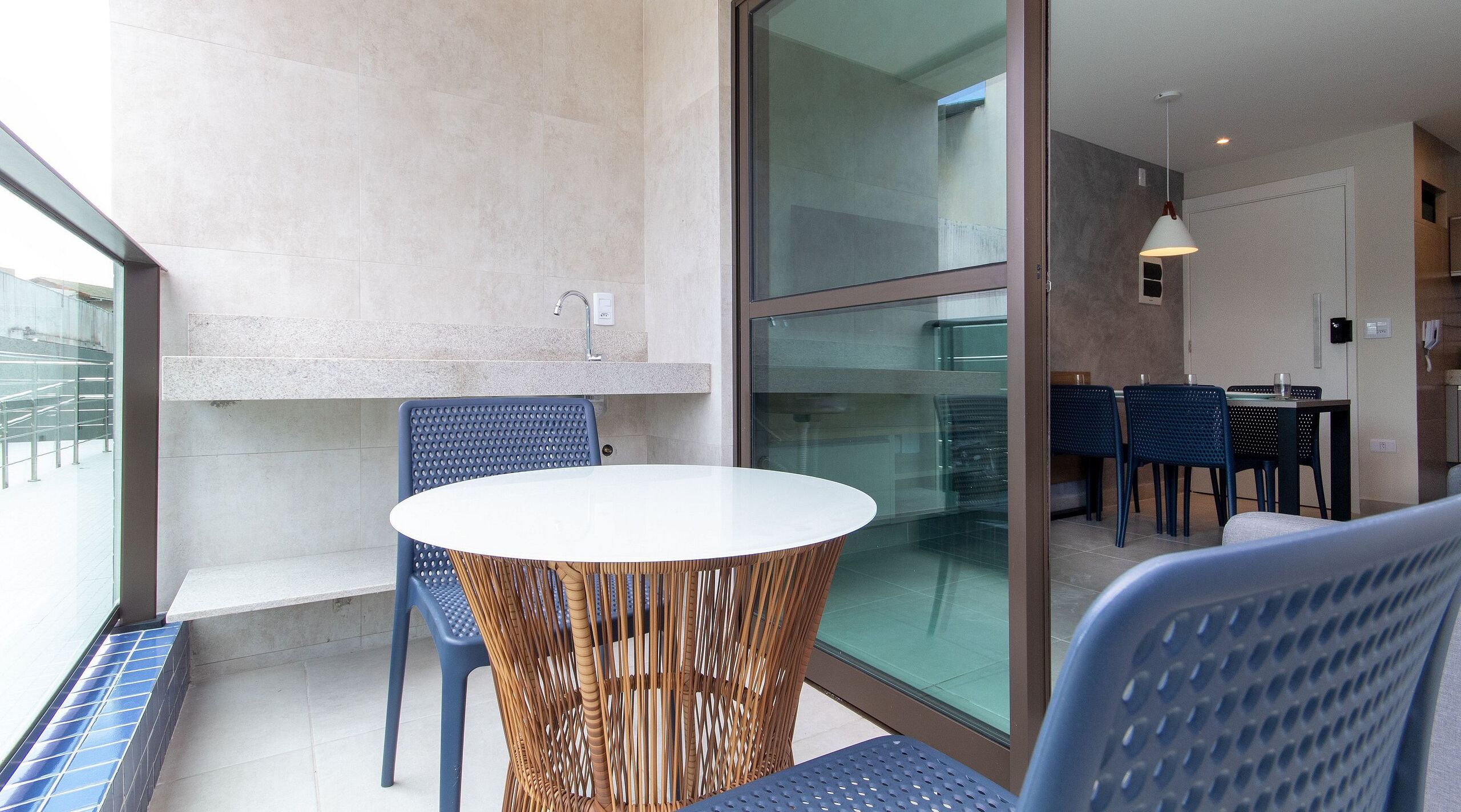 Uma varanda com mesa e cadeiras redondas, porta de vidro que dá para uma área interna com mesa de jantar e cadeiras, e vista para o exterior.