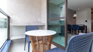 Een balkon met een ronde tafel en stoelen, een glazen deur die leidt naar een binnenruimte met een eettafel en stoelen, en uitzicht op buiten.