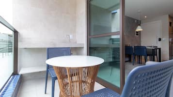 Uma varanda com mesa e cadeiras redondas, porta de vidro que dá para uma área interna com mesa de jantar e cadeiras, e vista para o exterior.