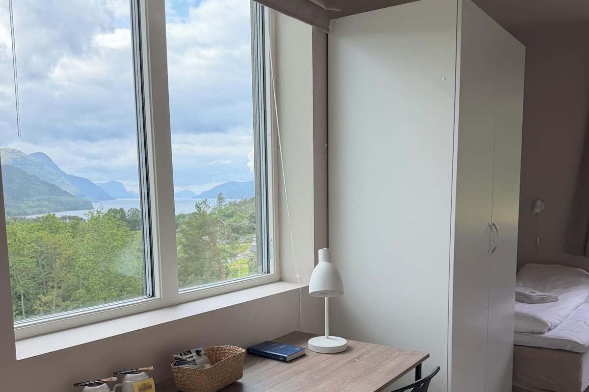 A bedroom with a bed, a desk with a chair, a lamp, and a view of mountains through the window.