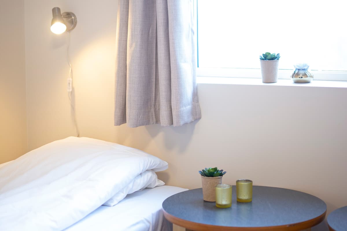 A small, neatly made bed with white linens, a wall-mounted light, a window with a curtain, and a small table with two candles and a potted plant.